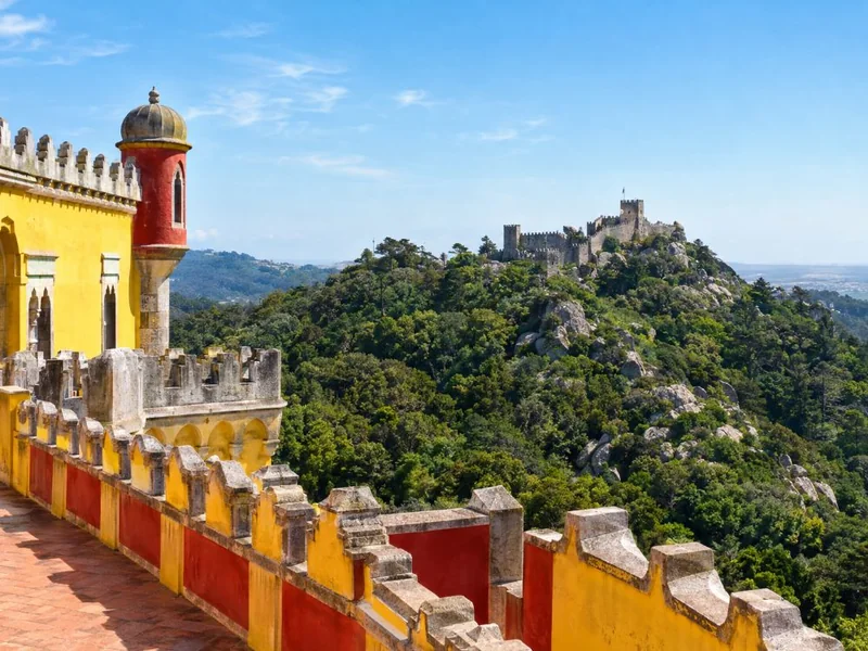 A red and yellow crenellated terrace looking across forested hills to grey castle walls on the next ridge