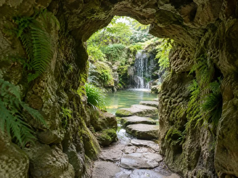 A rough stone grotto tunnel opening onto a small lake with stepping stones and a waterfall