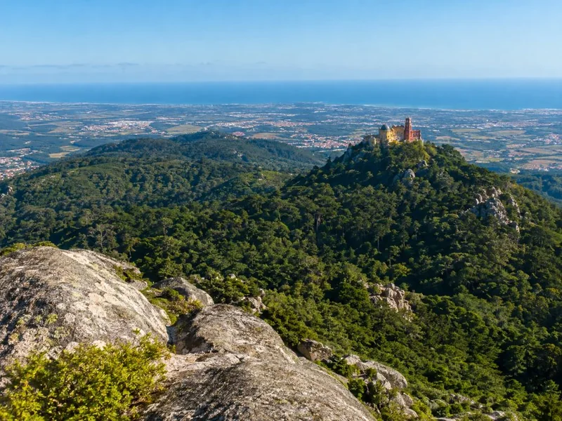 A granite lookout over green forested hills with a colourful palace on a distant peak and the ocean beyond